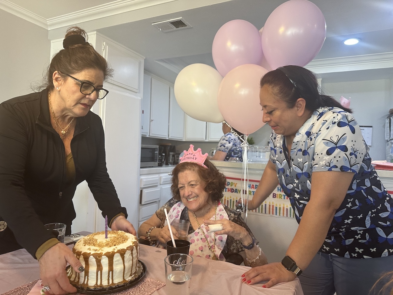 Three women celebrating a birthday with balloons and cake in a kitchen.
