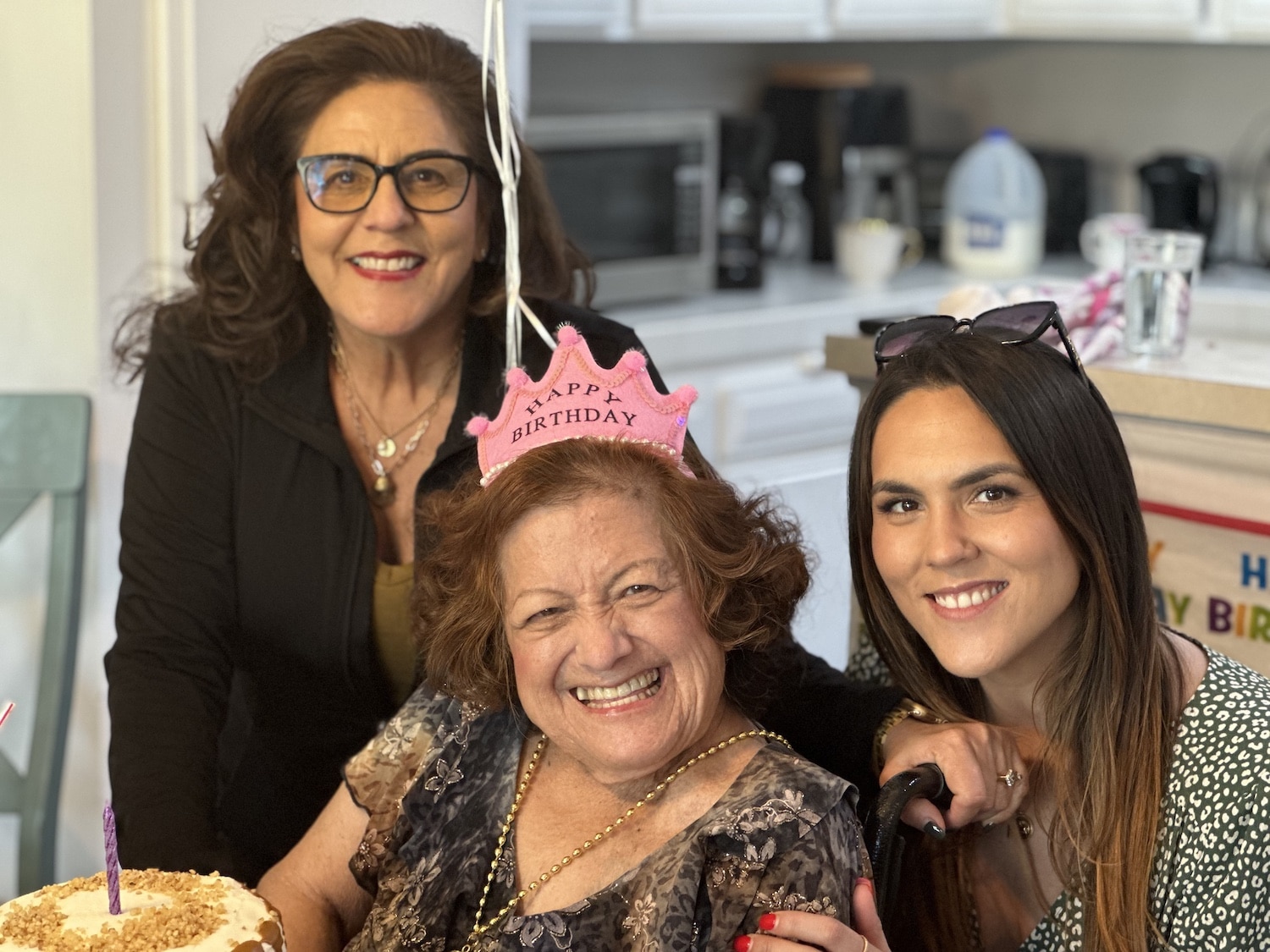 Three women celebrating a birthday with a cake and a Happy Birthday tiara indoors.