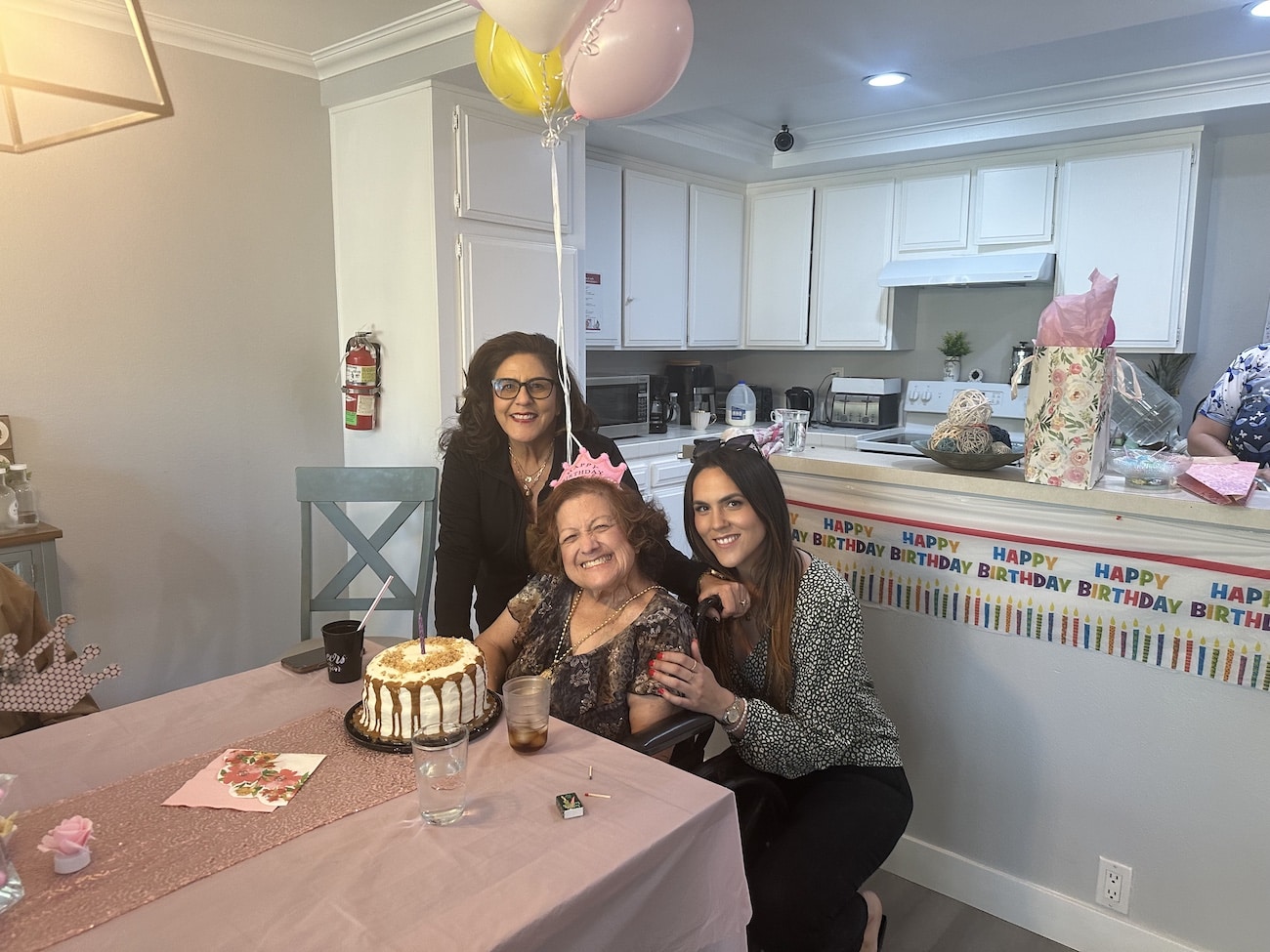 Three women celebrating a birthday with a cake and balloons in a decorated kitchen.