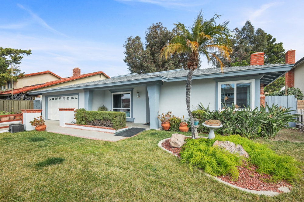 Suburban house with a well-kept lawn, palm tree, and a gray roof under a clear blue sky.