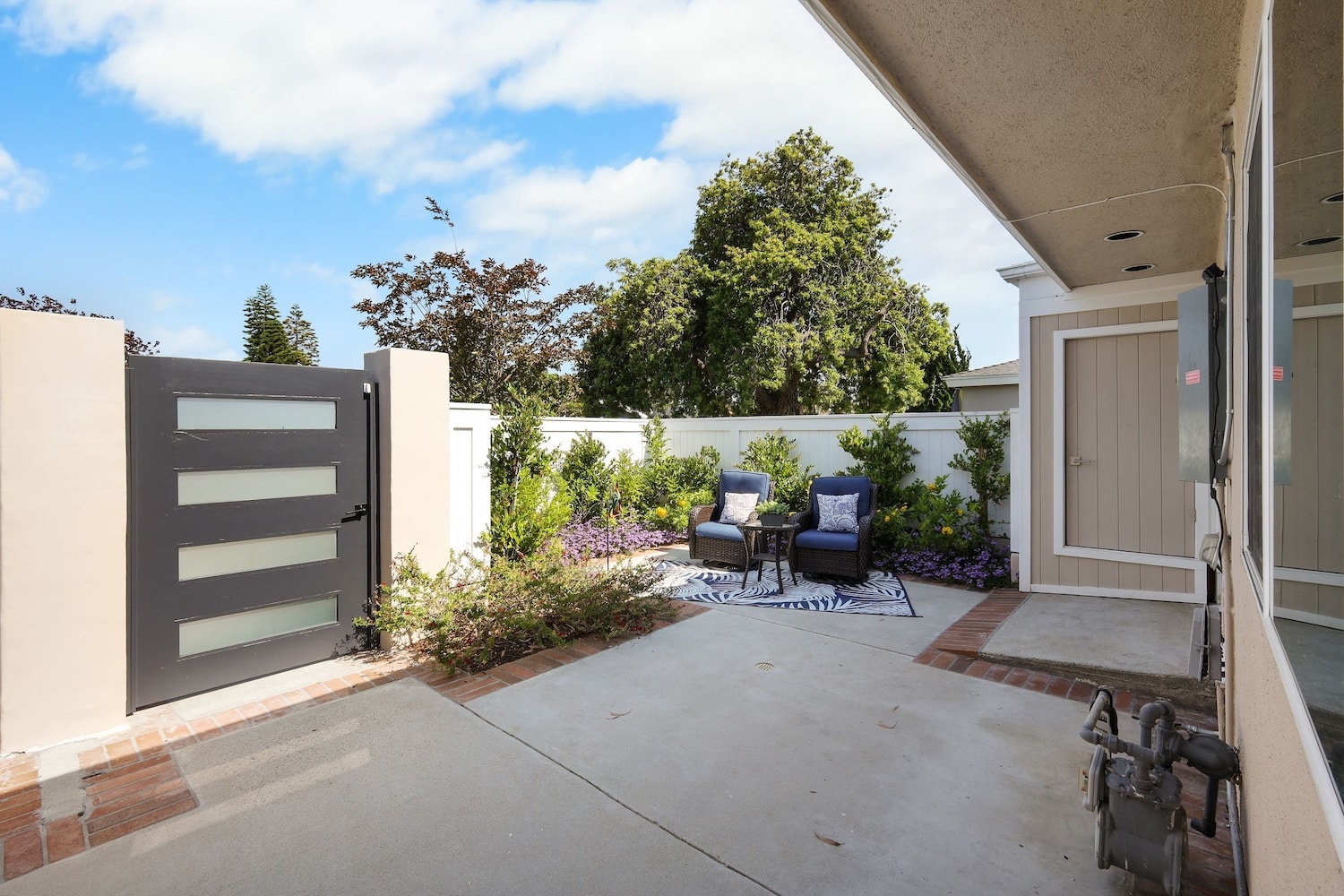 Modern patio with two blue chairs, a table, and a garden view under blue skies.