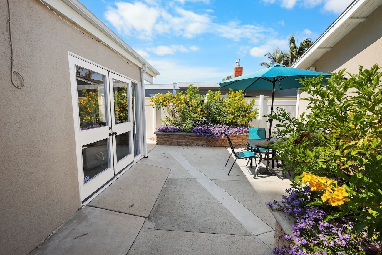 Sunny patio with colorful flowers, a table, chairs, and a teal umbrella against a blue sky.