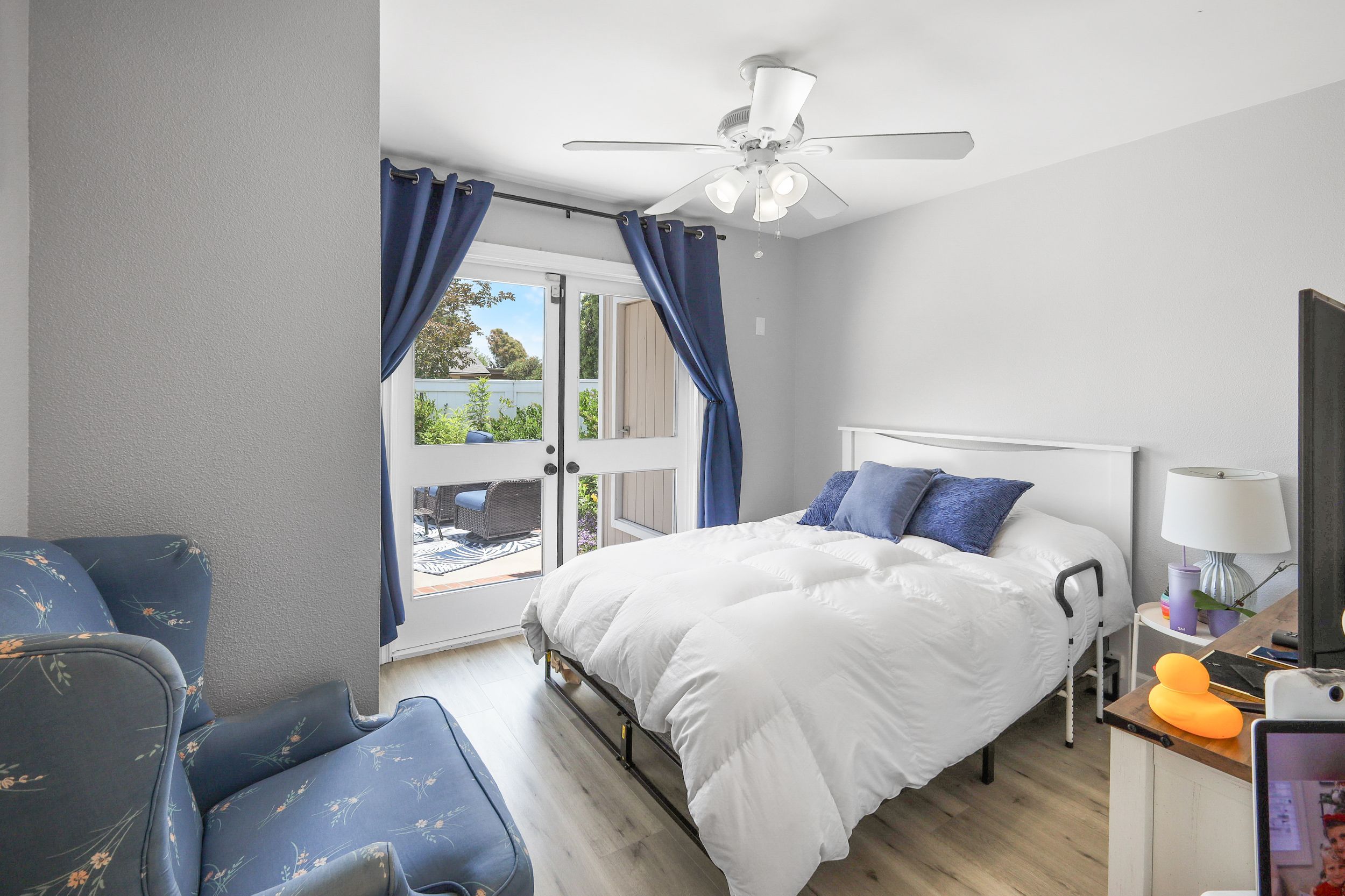 Bright bedroom with white bed, blue pillows, floral chair, ceiling fan, and French doors leading to patio.