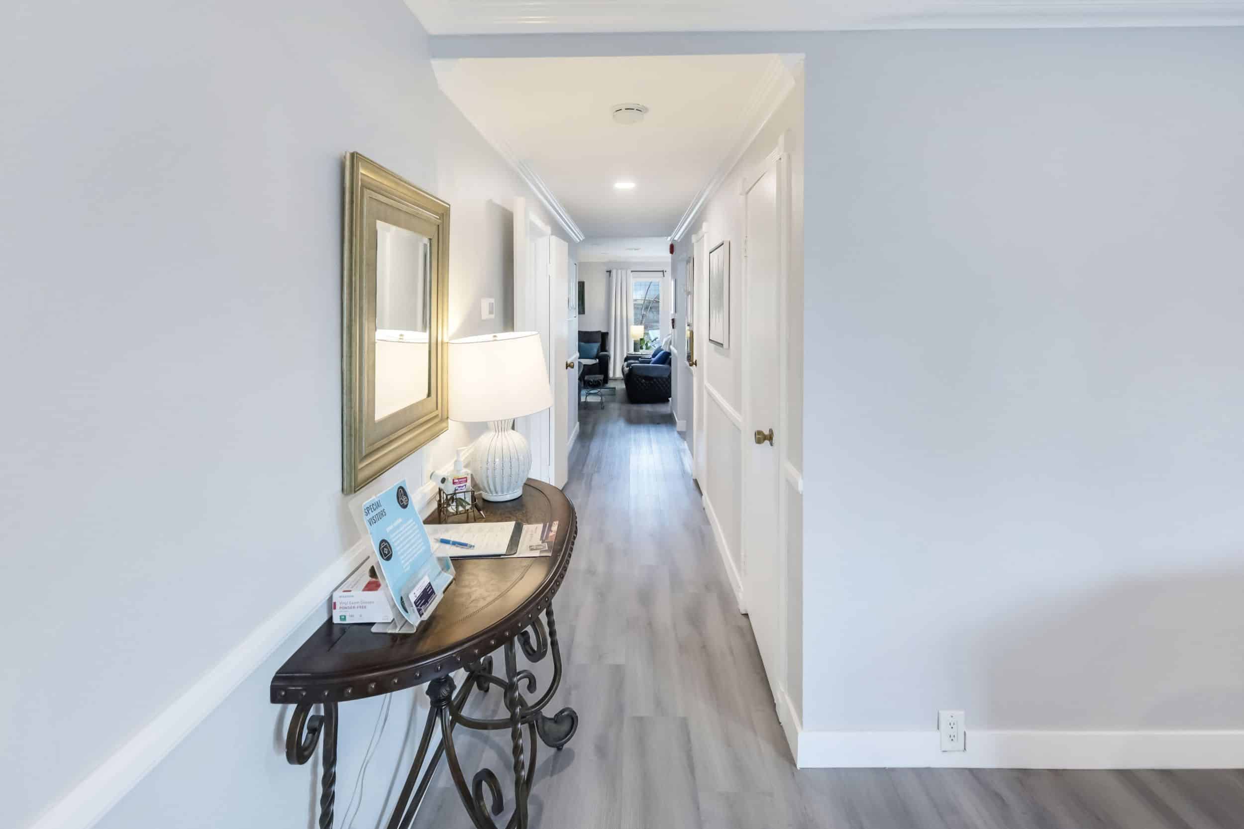 Bright hallway with table, lamp, framed artwork, and wood flooring leading to a living room in the distance.