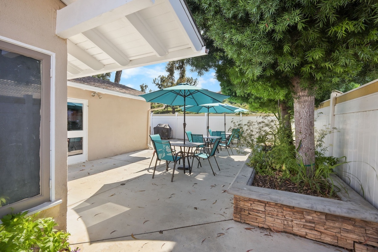 Outdoor patio with tables, chairs, and teal umbrellas, surrounded by greenery and fence.