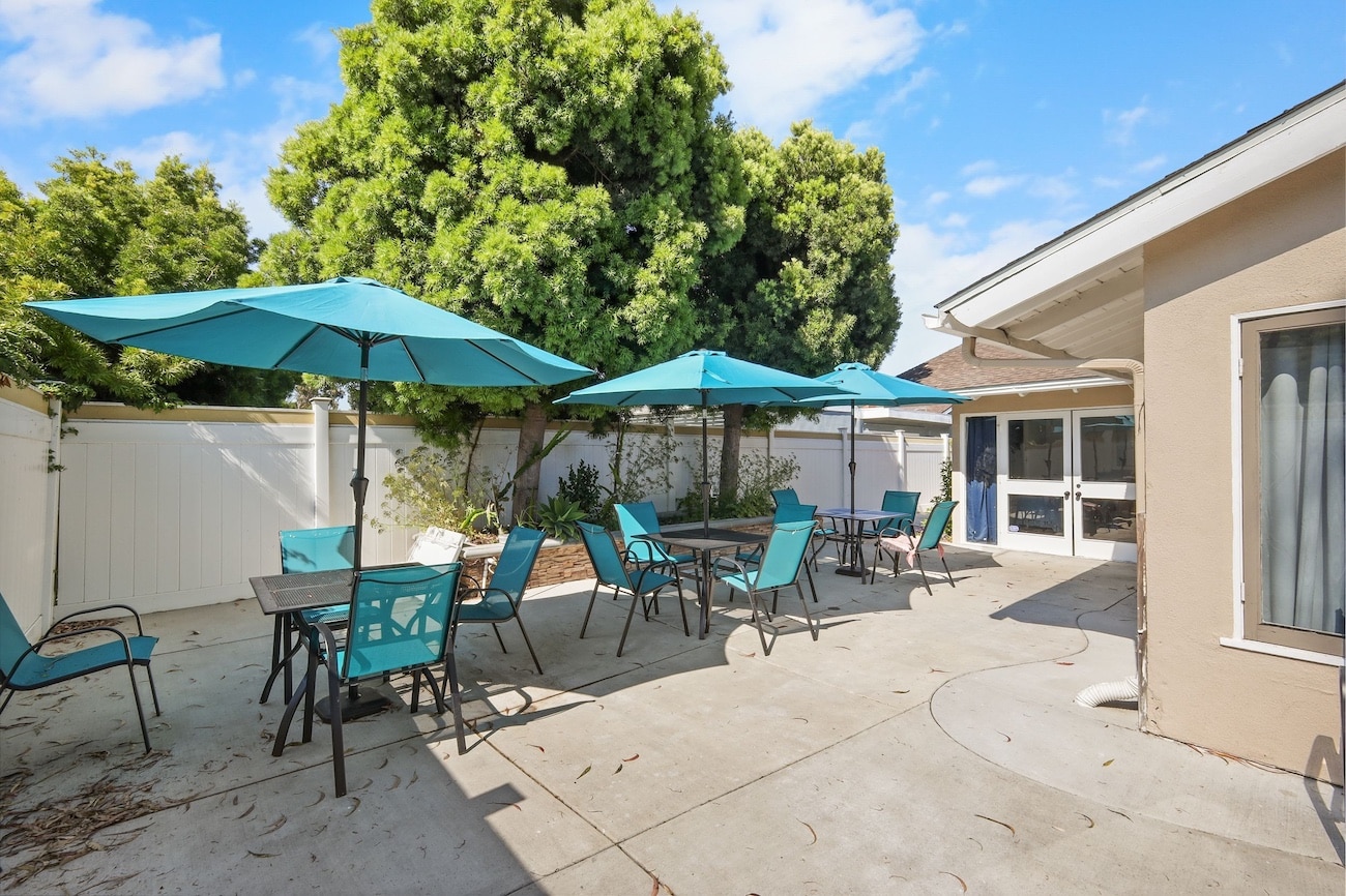 Outdoor patio with turquoise umbrellas and chairs, surrounded by green trees and a white fence.