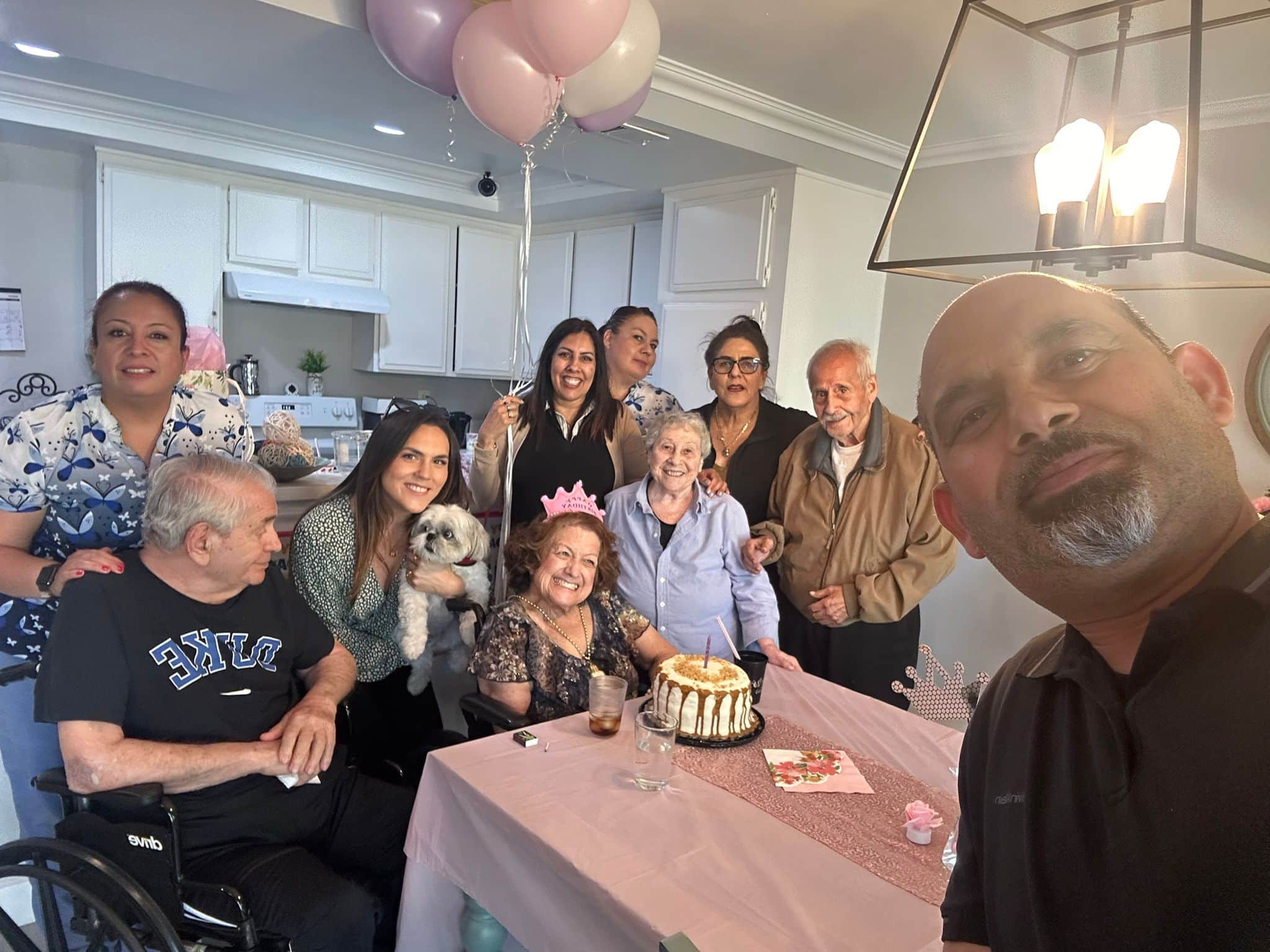 Family celebrating a birthday with cake and balloons indoors, including elderly members and a small dog.