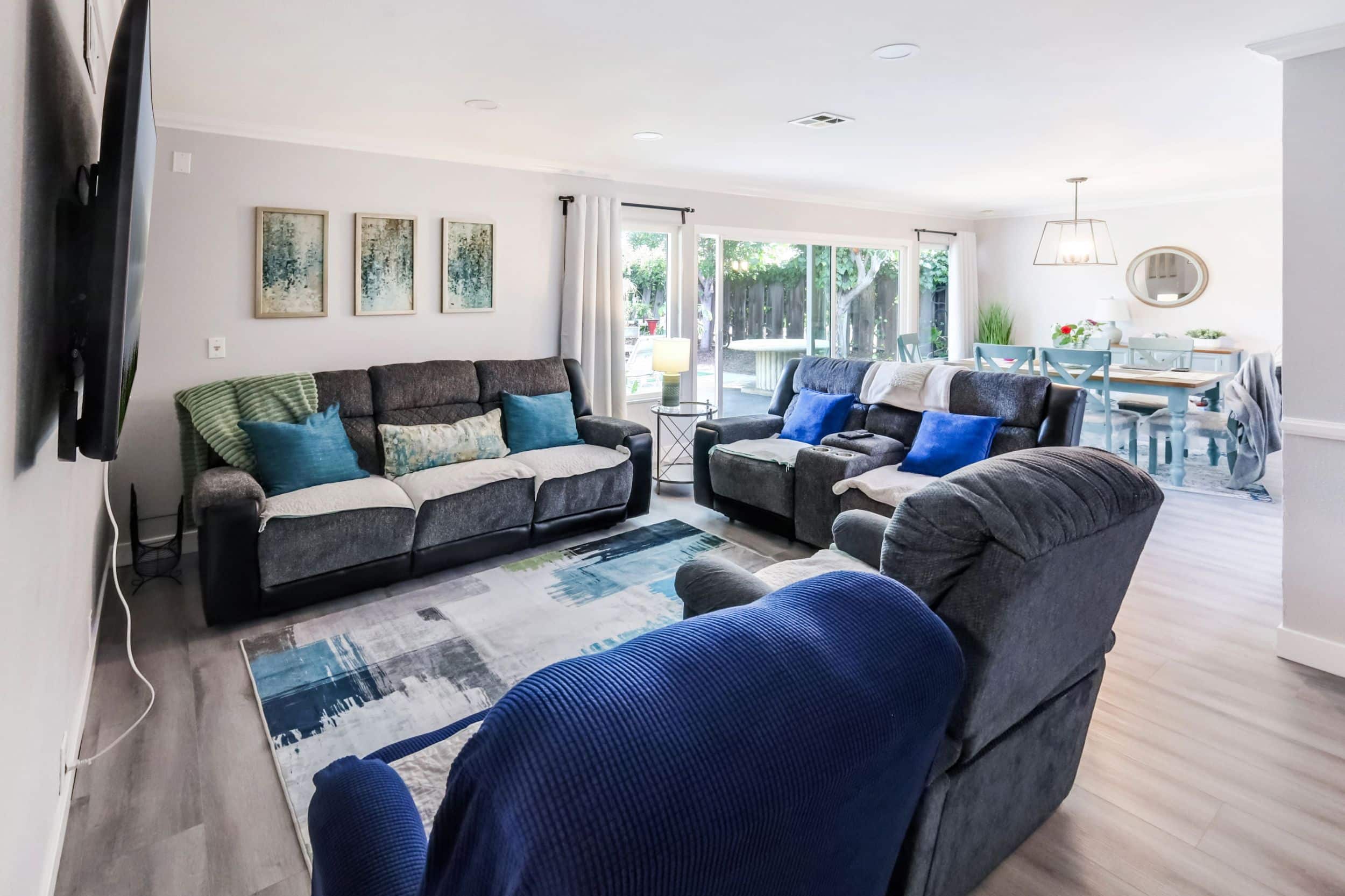 Modern living room with sofas, blue cushions, large windows, and a dining area with wooden table and chairs.