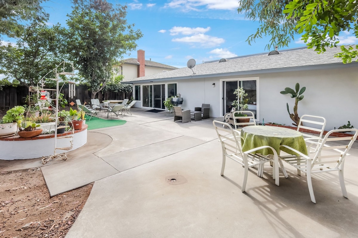 Spacious backyard patio with tables, chairs, potted plants, and trees under a clear blue sky.