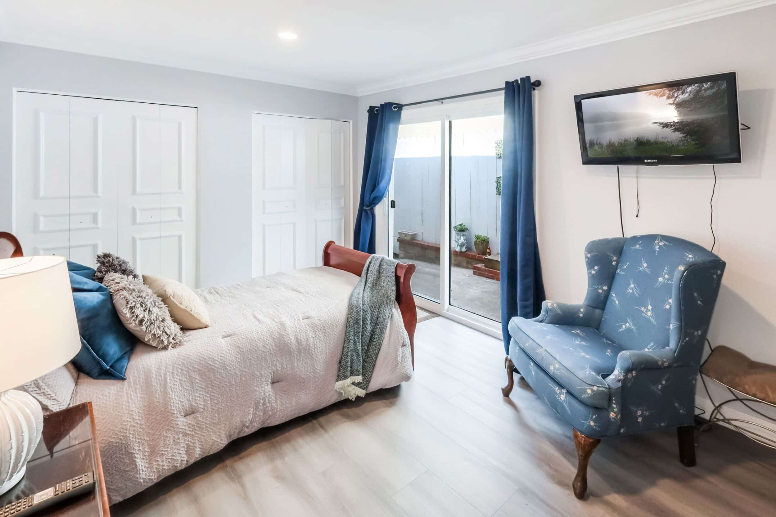 Cozy bedroom with blue chair, sliding glass door, and TV on wall. Bed with pillows and blanket.