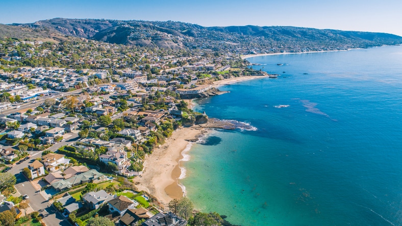 Aerial view of coastal town with sandy beach, clear blue ocean, and residential homes on hillside.