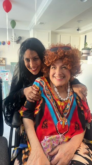 Two smiling women in a colorful living room, one sitting and the other standing behind her.
