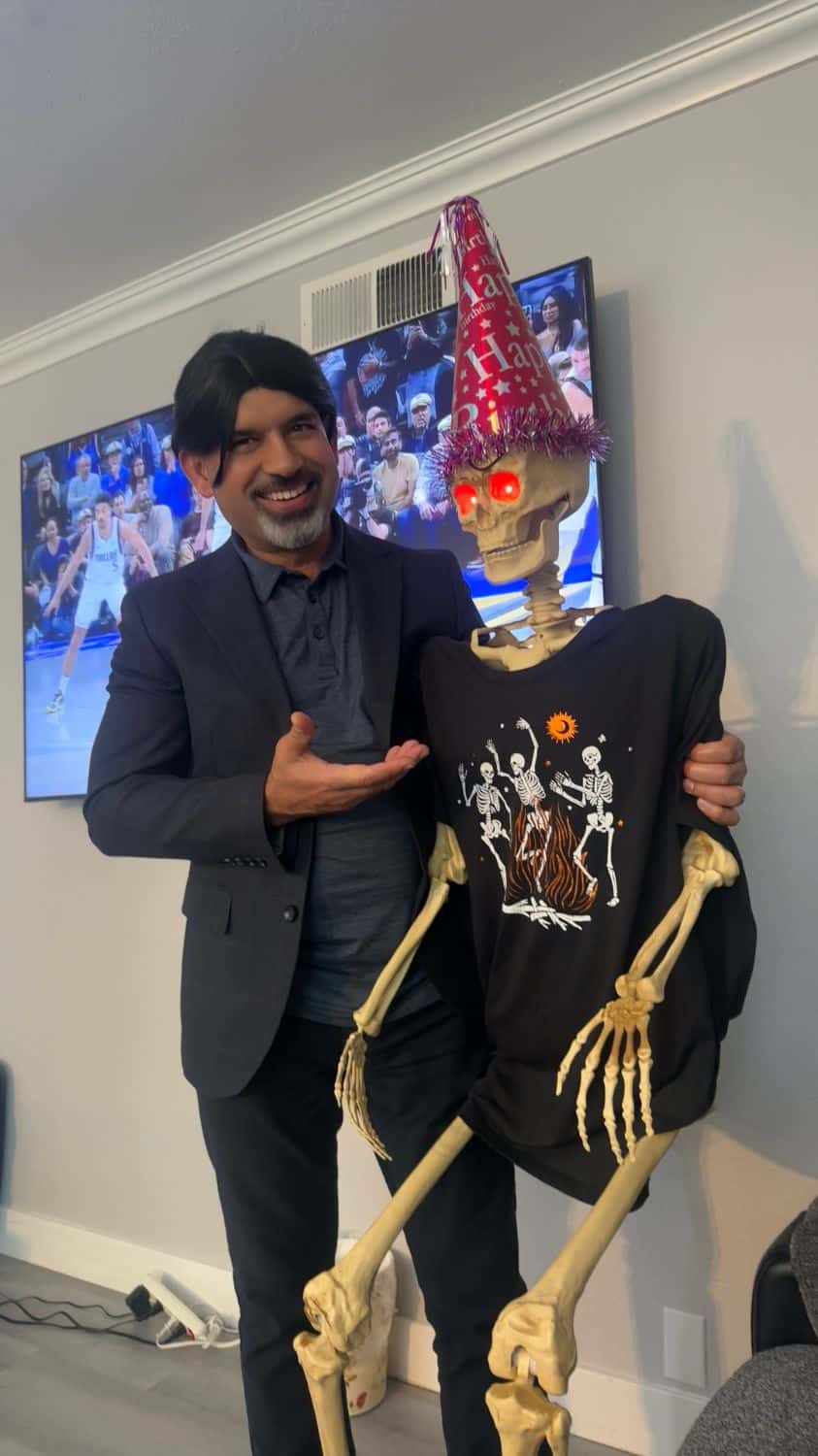 Man smiling with a skeleton wearing a party hat and shirt indoors.
