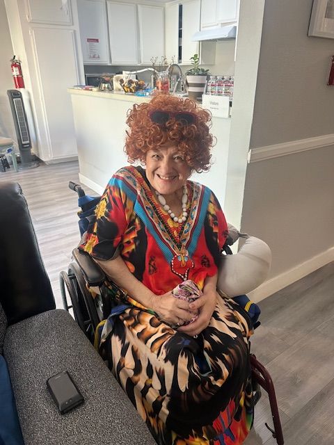 A person in colorful outfit smiles while sitting in a wheelchair in a cozy room.
